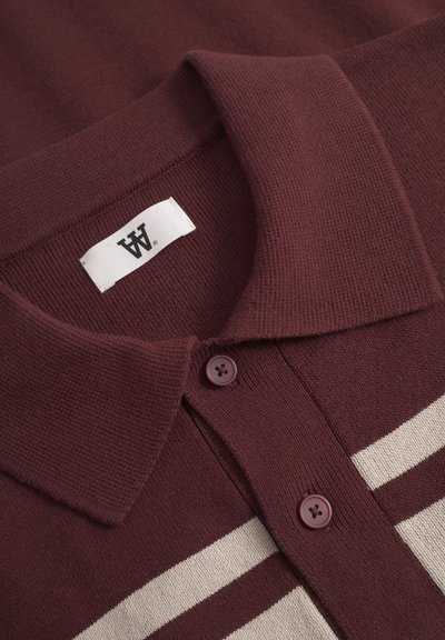 Gros plan sur le col d'une chemise en tricot bordeaux avec deux boutons et une étiquette blanche, présentant deux rayures horizontales crème sur le tissu.
