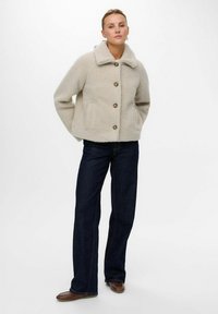 Veste en polaire beige avec un col montant, cinq boutons sur le devant et deux poches latérales. Associée à un jean en denim foncé et des chaussures marron.