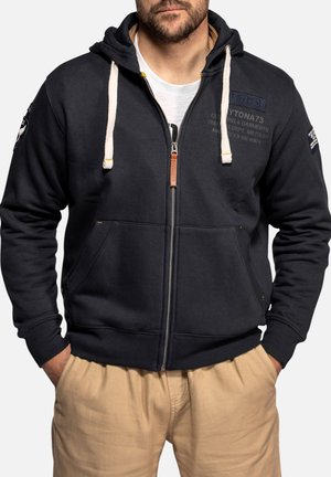 Homme portant un sweat à capuche noir zippé avec des cordons blancs, pantalon beige et T-shirt blanc, mains dans les poches, face à nous.