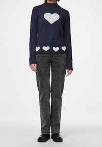 Pull bleu marine avec un motif de cœur blanc au centre et de petits cœurs le long de l'ourlet, associé à un pantalon en jean gris et des chaussures noires.