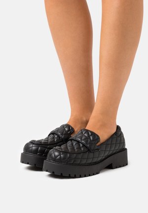 Mocassini neri trapuntati con suola chunky, caratterizzati da un prominente motivo a fiocco e superficie testurizzata. Ideali per un abbigliamento casual o smart-casual.