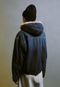 Jeanjacka med huva, med en foder av fuskpäls, mörkblå färg, elastisk nederkant och synliga söm detaljer. Matchad med grå byxor.