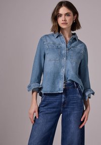 Hellblaues Jeanshemd mit zwei Brusttaschen, Knopfleiste, hochrollbaren Ärmeln und einem lockeren Schnitt. Kombiniert mit dunkelblauen, hochgeschnittenen Jeans.