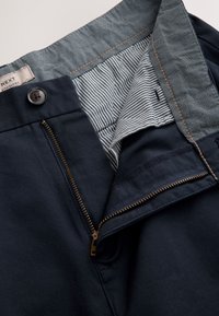 Pantalon bleu marine avec doublure intérieure rayée, fermeture éclair en laiton partiellement ouverte, bouton fermé et détail de ceinture cousu.