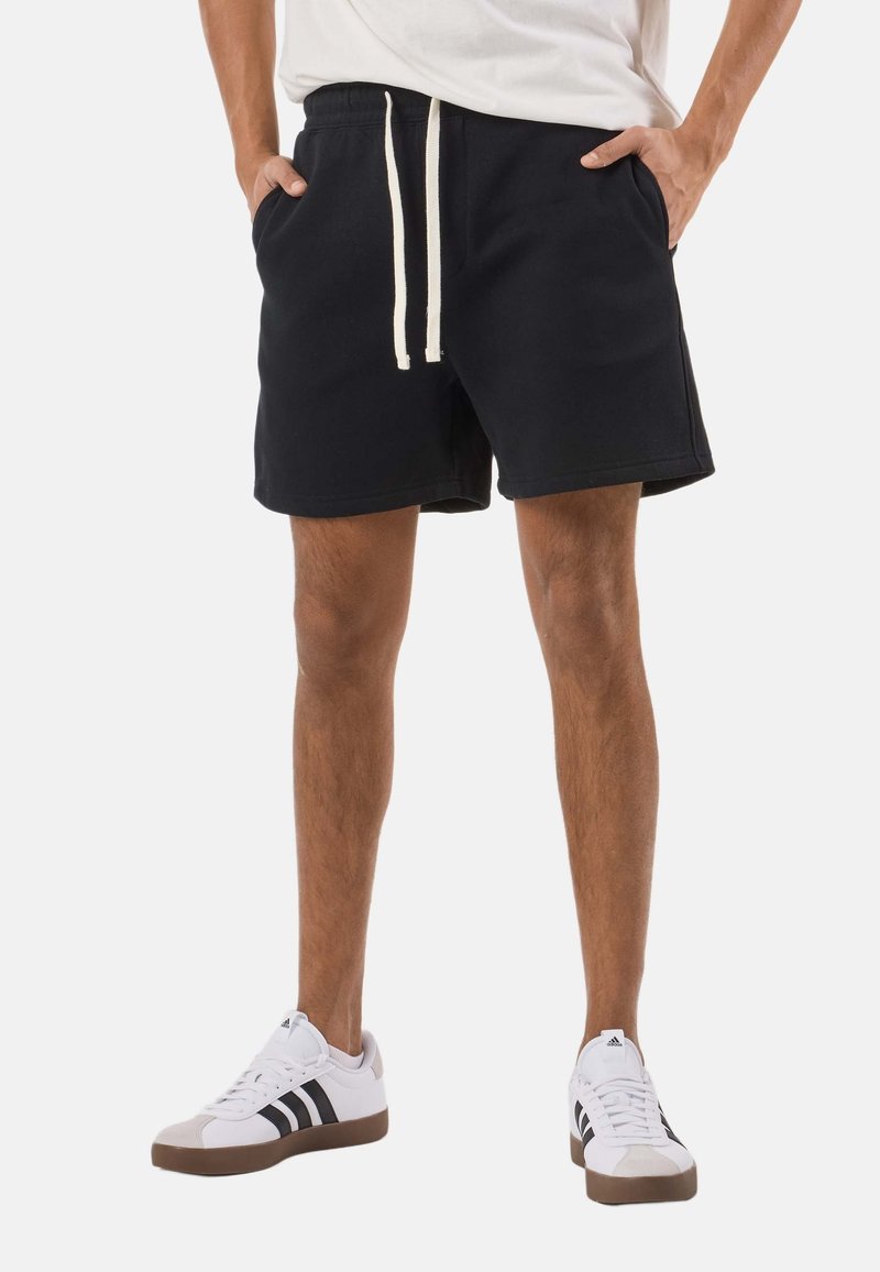 Shorts en coton noirs avec une ceinture élastique et un cordon de serrage blanc, dotés de poches latérales et d'une coupe décontractée, assortis à des baskets blanches.