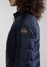Marineblauwe donsjacket met een quilted design en een ronde logo patch op de mouw. Voorzien van een ritssluiting en een verstelbare kraag.