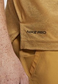 Nike Pro Sportbekleidung, goldbrauner Stoff, strukturierte Oberfläche, mit schwarzem Logo, einer Taschenakzentuierung und gerippten Details.
