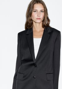 Černé sako na míru s výstřihem s vykrojením, zapínáním na jeden knoflík a dvěma předními kapsami. Hladká látka s dobře tvarovaným střihem.