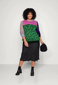 Usmievajúca sa žena s kučeravými vlasmi má na sebe sveter s farebnými blokmi a zeleným leopardím vzorom, čiernu midi sukňu, čierne lodičky a drží nadýchanú čiernu tašku.