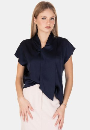 Donna che indossa una blusa blu navy a maniche corte con un dettaglio a nodo davanti e pantaloni chiari a vita alta, in piedi contro uno sfondo semplice.