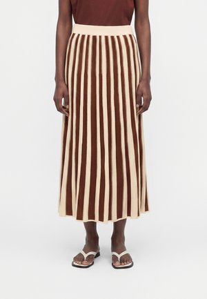 Personne portant une jupe midi rayée verticale marron et beige, un haut marron et des sandales blanches, debout devant un fond clair.