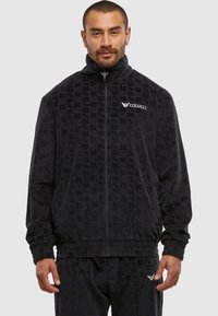 Homme portant une veste zippée noire texturée et un pantalon assorti avec le logo Colucci sur la poitrine et le pantalon, sur un fond uni.