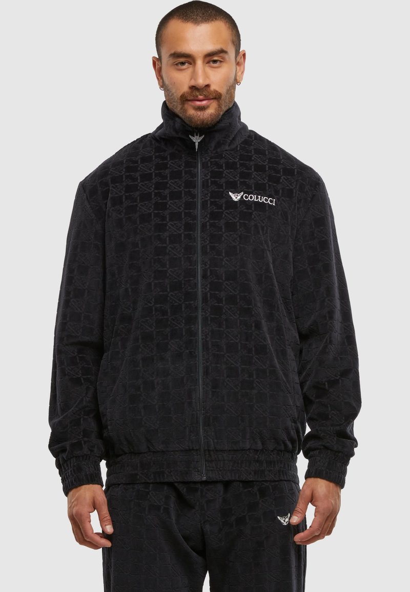 Homme portant une veste zippée noire texturée et un pantalon assorti avec le logo Colucci sur la poitrine et le pantalon, sur un fond uni.