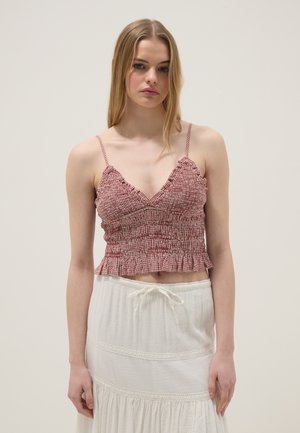 Jonge vrouw met lang blond haar, gekleed in een rood patroon crop top en een witte rok met trekkoord, staand tegen een effen achtergrond.