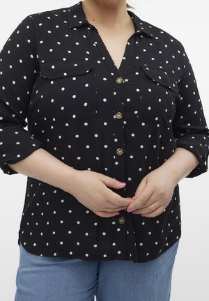 Osoba ubrana w czarną koszulę na guziki z białymi kropkami i podwiniętymi rękawami, w jasnoniebieskich dżinsach, z rękami złączonymi przed sobą.