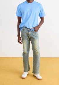 Ljusblå bomulls-T-shirt med en liten logotyp, kombinerad med bleknade blå jeans och vita sneakers med en beige sula. Enfärgad bakgrund.