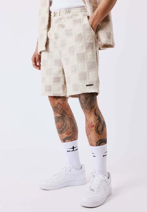 Homme portant un short beige texturé, jambes tatouées, chaussettes blanches avec motif noir et baskets blanches sur un fond uni.