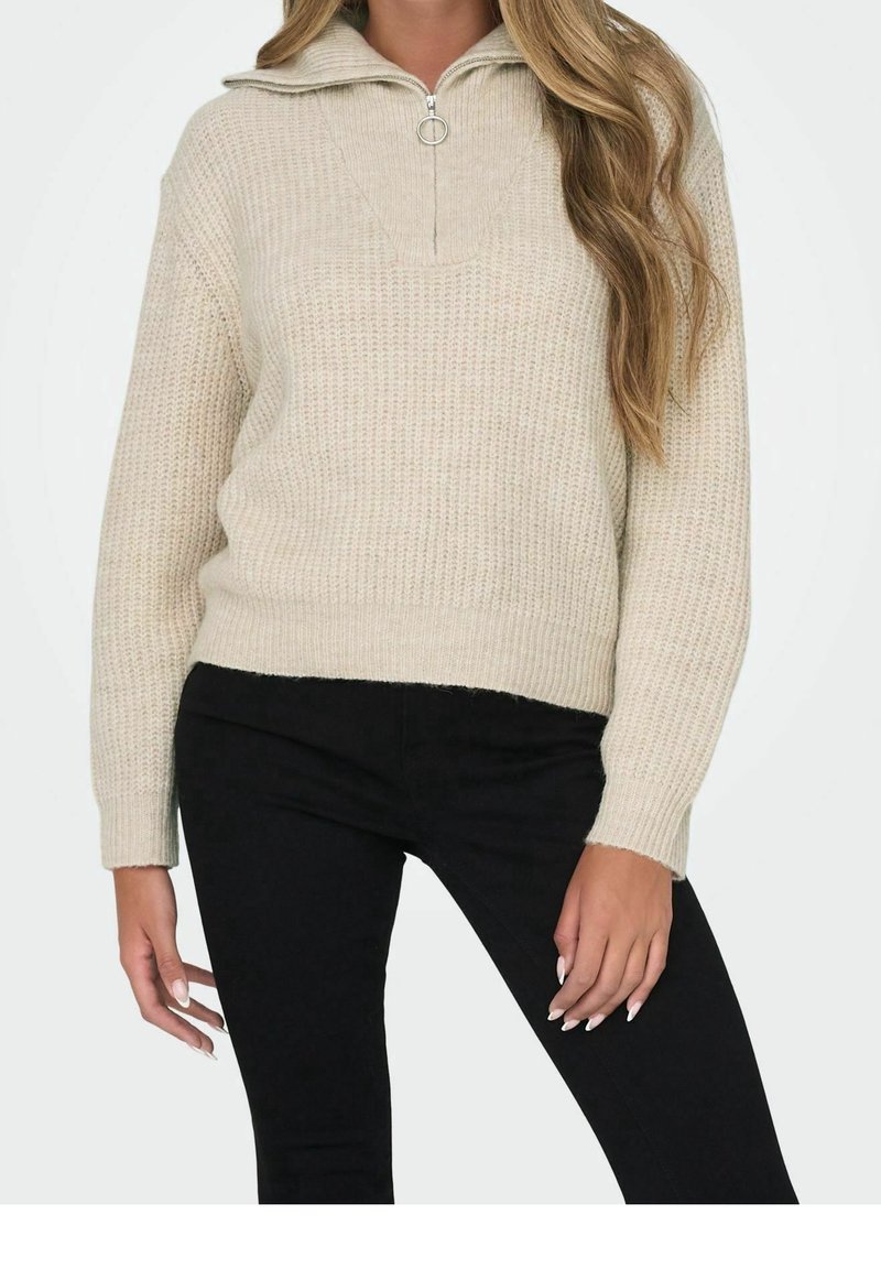 Femme portant un pull en maille beige avec un col à fermeture éclair partielle et un pantalon noir, debout avec une main près de sa cuisse et l'autre détendue.