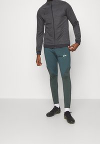 Pánský atletický outfit skládající se z černé texturované zipové bundy a tyrkysových zúžených kalhot s geometrickými vzory, doplněný černými kopačkami.