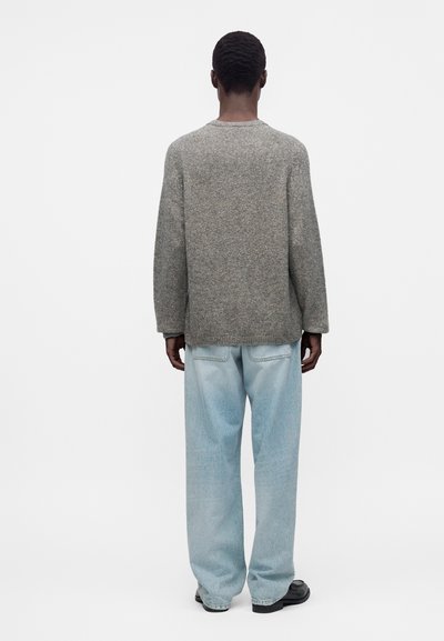Personne debout de dos, portant un pull gris à manches longues, un jean large bleu clair et des chaussures noires sur un fond blanc uni.