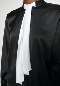 Blusa in satin nero con dettaglio in cravatta bianca sul collo. La blusa ha un colletto ed è liscia al tatto con un aspetto aderente.