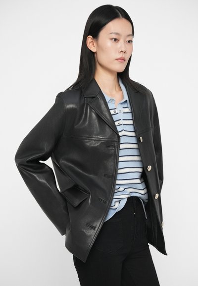 Γυναίκα με ίσια μαύρα μαλλιά, φορώντας μια μαύρη δερμάτινη σακάκι με κουμπιά πάνω από ένα ανοιχτό μπλε πουκάμισο με ρίγες και μαύρο παντελόνι, με τα χέρια πίσω από την πλάτη.