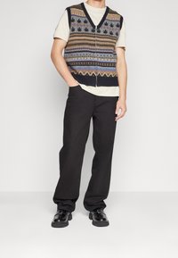 Pletená vesta s výstřihem do V v tmavých barvách, zdobená geometrickými vzory. Nositelná přes světlé tričko, doplněná černými širokými kalhotami a botami.