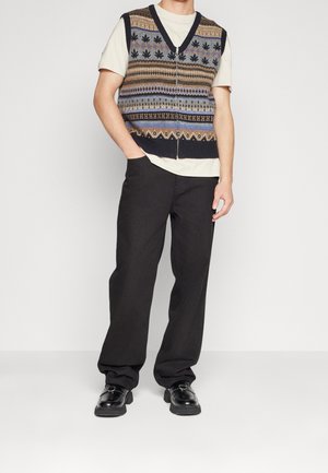 Sweter bez rękawów z głębokim dekoltem w ciemnych kolorach, ozdobiony geometrycznymi wzorami. Noszony na jasnej koszulce, w połączeniu z czarnymi spodniami o szerokich nogawkach i butami.