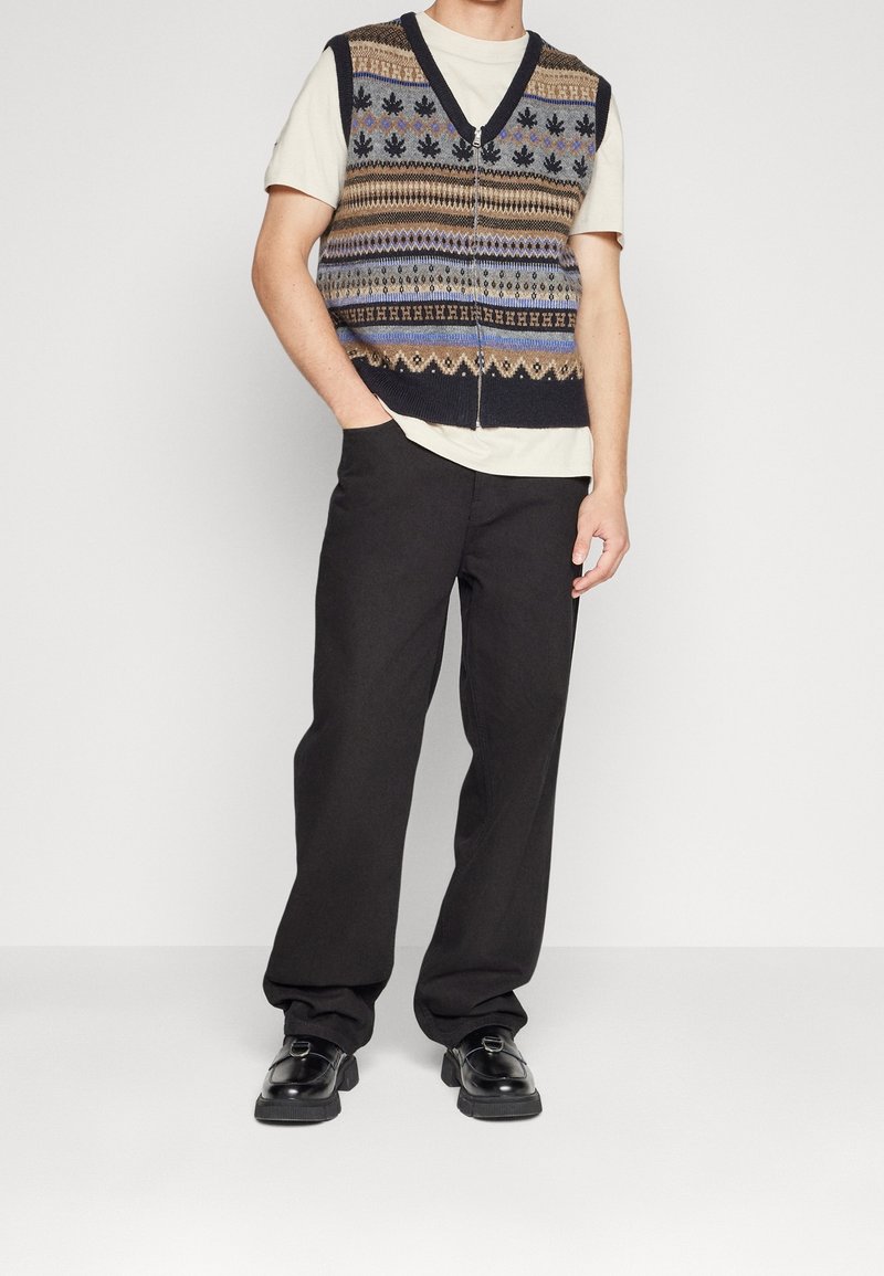 Pletená vesta s výstřihem do V v tmavých barvách, zdobená geometrickými vzory. Nositelná přes světlé tričko, doplněná černými širokými kalhotami a botami.