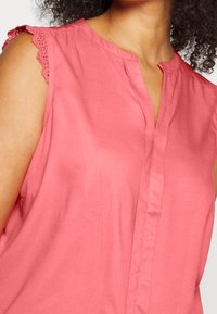 Różowa bluzka bez rękawów o miękkiej fakturze, z dekoltem w kształcie litery V i delikatnymi koronkowymi akcentami na ramionach. Gładkie wykończenie tkaniny.