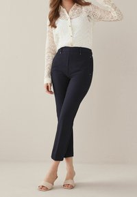 Donna che indossa una blusa bianca in pizzo a maniche lunghe, pantaloni neri aderenti alla caviglia e sandali aperti beige, in piedi all'interno.