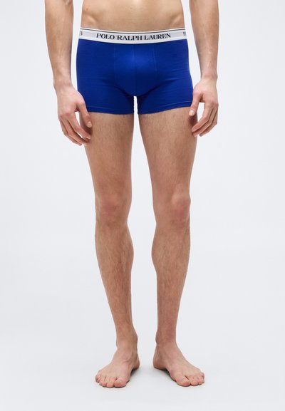 Αρσενικός κορμός και πόδια που φοράνε μπλε boxer briefs Polo Ralph Lauren με λευκή ελαστική ζώνη με λογότυπο, όρθιος ξυπόλυτος σε απλό φόντο.