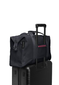 Sac de sport en tissu durable de couleur marine, équipé de poignées doubles, d'un dessus zippé et d'une poche extérieure avec une fermeture éclair rose, posé sur une valise noire.