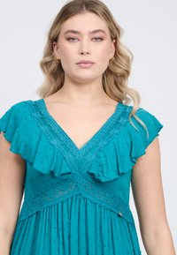 Vestido verde azulado con escote en V, mangas con volantes y detalles de encaje en la cintura. Tela texturizada con pequeños puntos en toda su superficie.