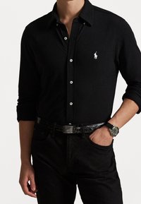 Zwart shirt met lange mouwen en een knoopkraag, voorzien van een wit logo. Gecombineerd met een zwarte broek en een horloge met een groene wijzerplaat en een zwarte band.