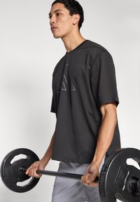 Μαύρο διχτυωτό αθλητικό μπλουζάκι με άνετη γραμμή και κοντά μανίκια, με γεωμετρικό σχέδιο στο στήθος, που κρατά ένα άτομο ενώ σηκώνει μπάρα με βάρη.