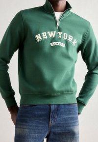 Zelená mikina na zip s potiskem "NEW YORK" a "SCHOTT" v bílé barvě. Hladká textura, žebrované manžety a lemy.