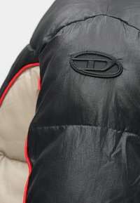 Juodas paminkštintas džemperis su blizgiu paviršiumi, turintis raudoną apdailą siūlėse ir juodą išsiuvinėtą logotipą ant peties.