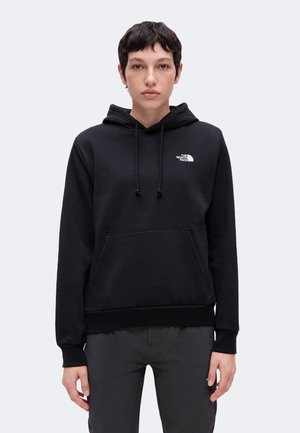 Persoon met kort donker haar, gekleed in een zwarte The North Face hoodie en donkere broek, staand tegen een effen lichte achtergrond.
