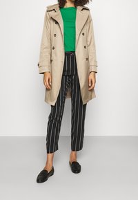 Beige trenchcoat med dubbelknäppning, grön skjorta under, svarta randiga byxor och svarta loafers, med en traditionell design.