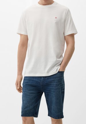 T-shirt en coton blanc avec col rond, manches courtes et petit logo rouge sur la poitrine, associé à un short en jean bleu.