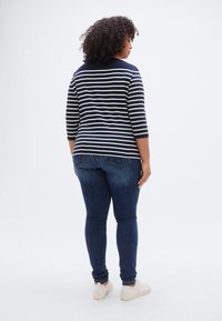 Μπλε ναυτικό και λευκό μπλουζάκι με μακριά μανίκια και στρογγυλή λαιμόκοψη, συνδυασμένο με σκουρόχρωμο στενό τζιν και λευκά αθλητικά παπούτσια, εμφανιζόμενο από πίσω.