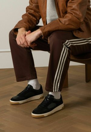 Zwart-witte sneakers met een gestructureerde suède bovenkant en rubberen zool, gecombineerd met een paarse trainingsbroek met witte strepen aan de zijkanten.