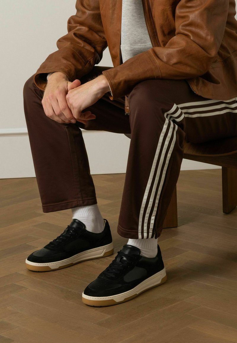 Zwart-witte sneakers met een gestructureerde suède bovenkant en rubberen zool, gecombineerd met een paarse trainingsbroek met witte strepen aan de zijkanten.