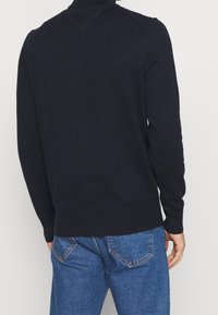 Uomo che indossa un maglione a collo alto blu scuro aderente e jeans di denim blu, visto da dietro su uno sfondo chiaro e semplice.