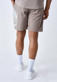 Pantalones cortos marrón claro con una textura suave, que presentan un gran diseño gráfico blanco en la pierna derecha y bolsillos traseros con broches metálicos.