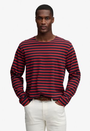 Hombre con una camisa de manga larga a rayas azul marino y rojo, pantalones blancos y un cinturón trenzado marrón, manos en los bolsillos.