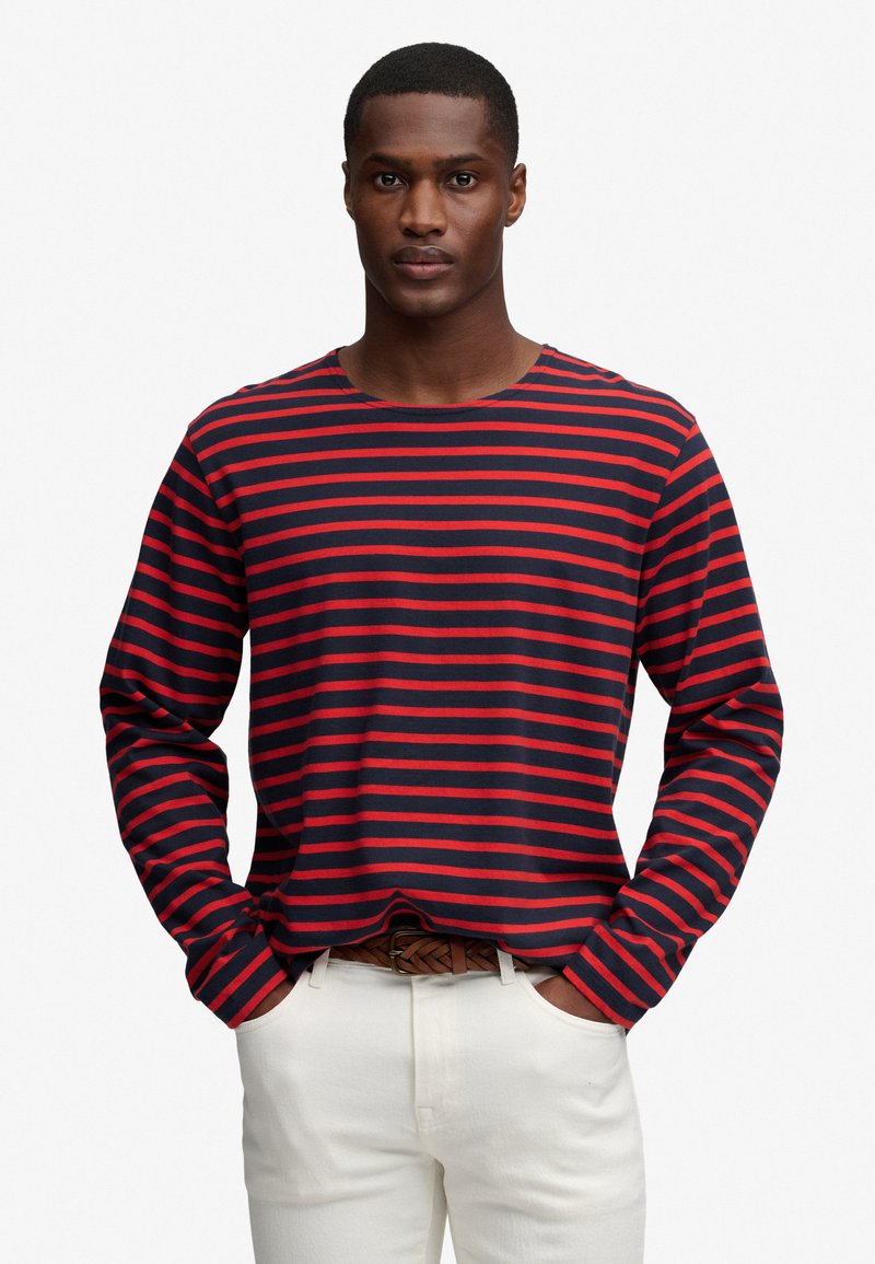 Homme portant une chemise à manches longues rayée bleu marine et rouge, pantalon blanc et ceinture tressée marron, mains dans les poches.