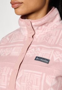 Lys rosa fleece-genser med halv-zip krage, med et geometrisk mønster, stoffknapper og en svart Columbia-logo-patch.