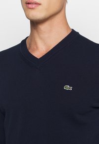 Suéter de punto azul marino con escote en V. Presenta un pequeño logo de cocodrilo verde en el área del pecho izquierdo.